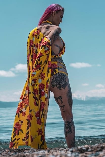 A woman on a beach, tying a vibrant tropical print Shein sarong around her waist as a skirt. She is wearing a bikini top and sunglasses, and the ocean is visible in the background.