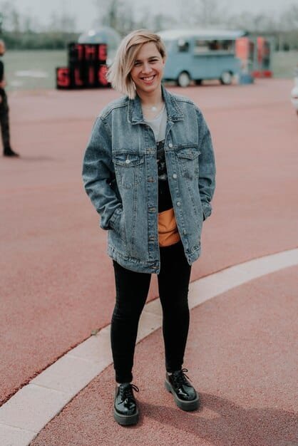 A woman stylishly layered in an oversized denim jacket over a hoodie, paired with leggings and combat boots. She's wearing a beanie and sunglasses, accentuating the casual, urban style. The background is an alley wall with street art.